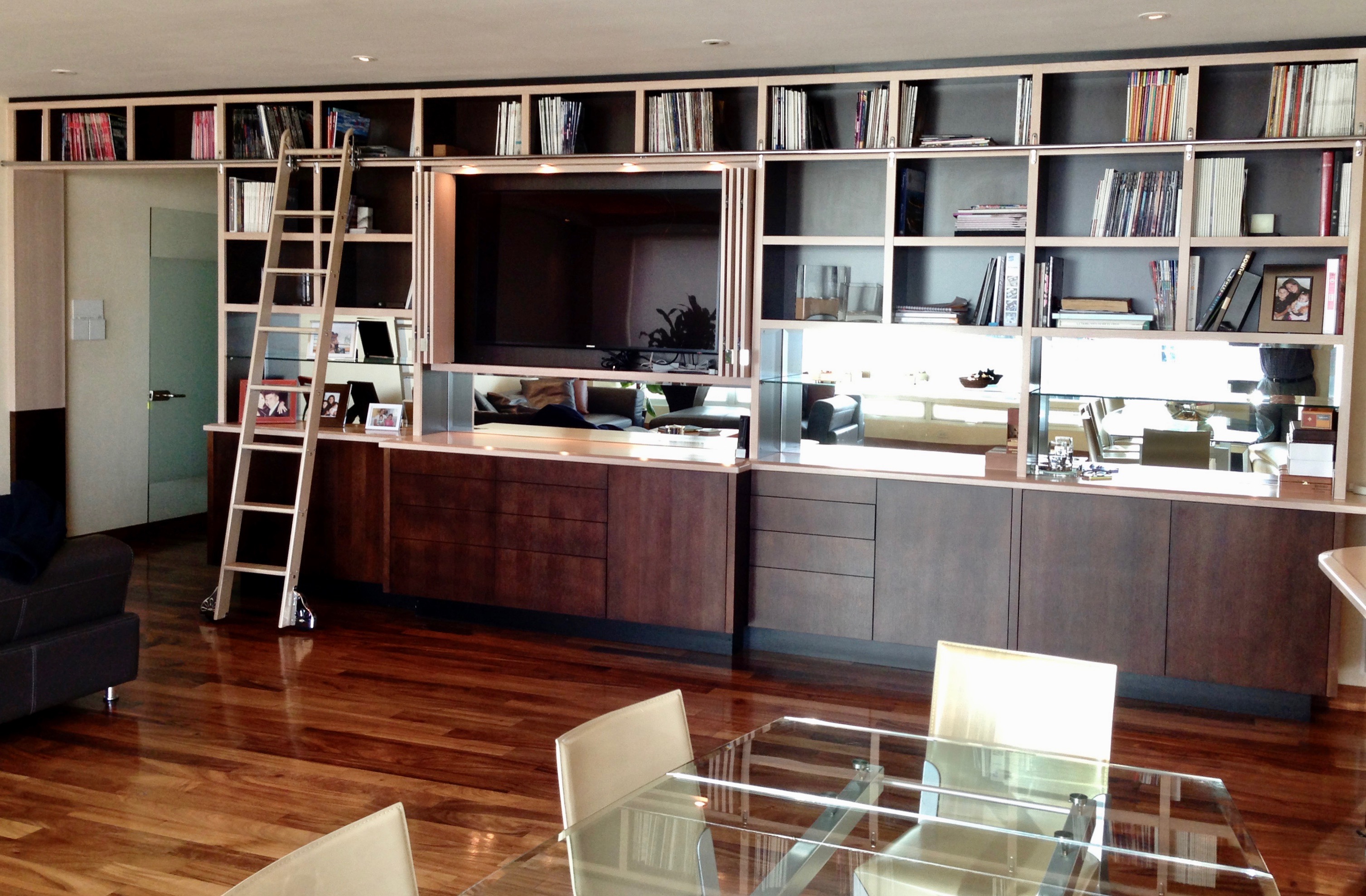 Custom home Libraries Display walls. A custom JerryJacobsDesign © library wall Saanta Fe high-rise, CDMX. Byfolds cover TV and show a custom mural, mirrored backsplash. Rollig ladder. Curved metamorphic glass table.